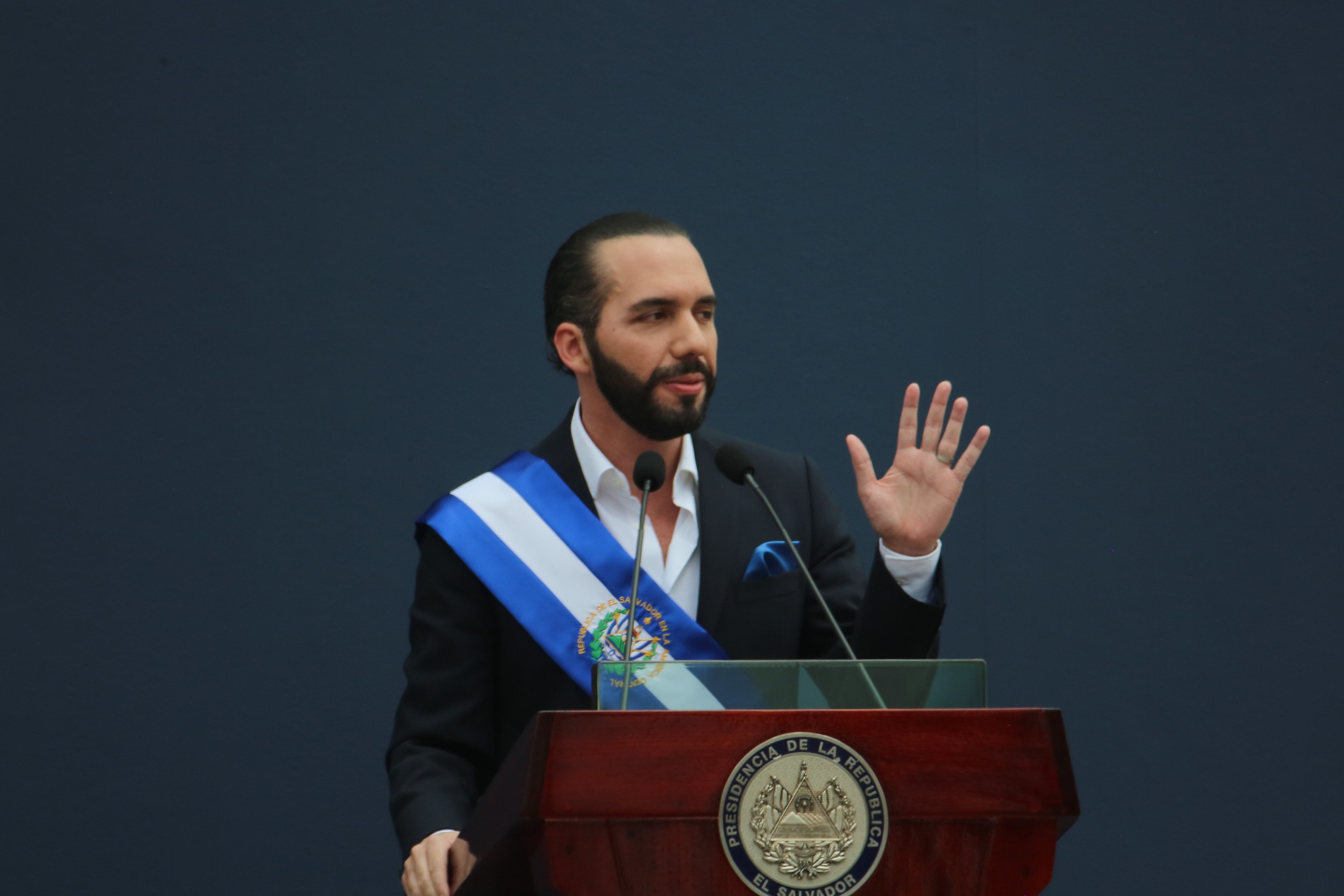 A man with a beard and slicked-back black hair speaks at a podium with his left hand raised in the air.