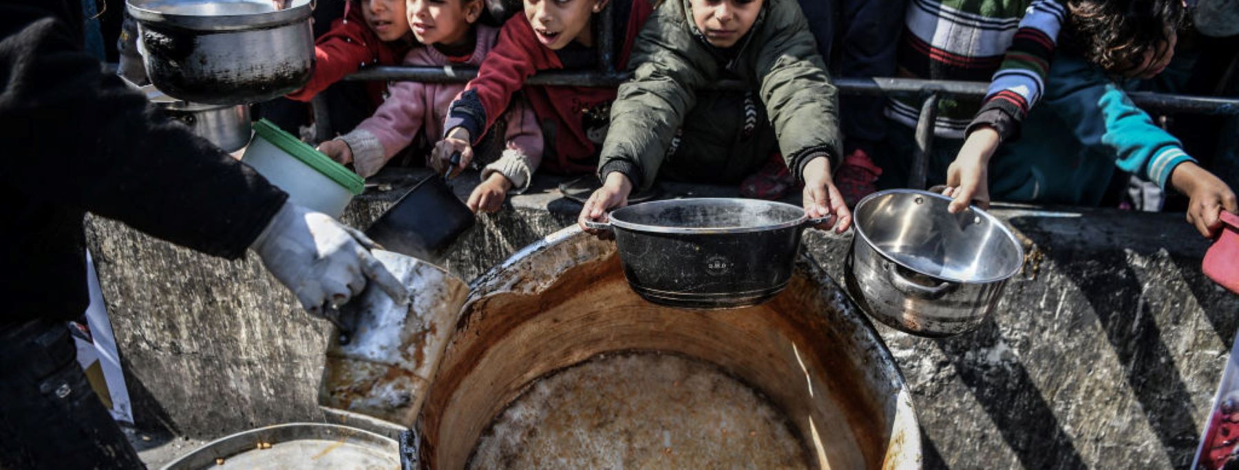 Palestinians hold out their empty containers to be filled with food, distributed by charity organizations, behind bars since they are unable to obtain basic food supplies due to the embargo imposed by Israeli forces in Rafah, Gaza on February 25, 2024. Photo by Abed Zagout/Anadolu via Getty Images.