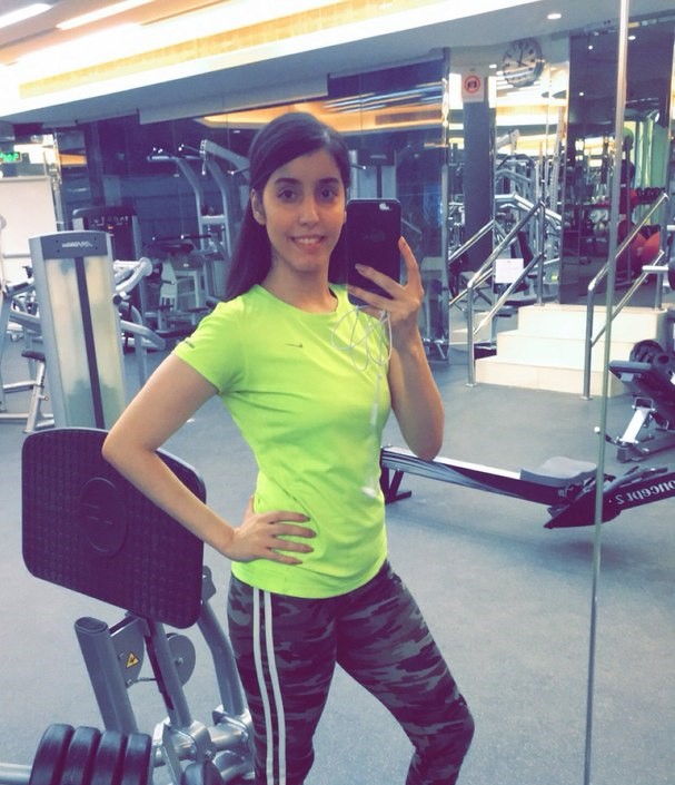 A picture of Manahel wearing a green short sleeve shirt and yoga pants. Her hair is in a ponytail and she appears to have taken the photo in a gym.