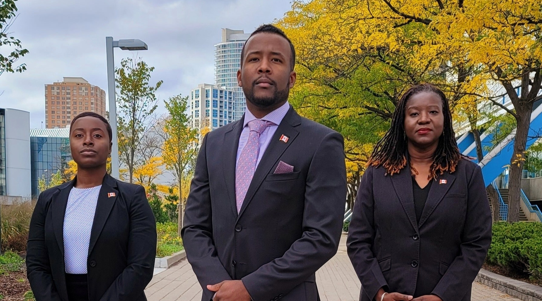 Two women and a man, each wearing a dark suit with a Canadian flag on the left lapel, stand outdoors on a brick walkway in autumn.