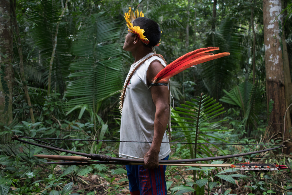 A man with wearing a crown of yellow feathers, a white tank top and shorts stands holding a bow and arrows in a densely wooded forest.