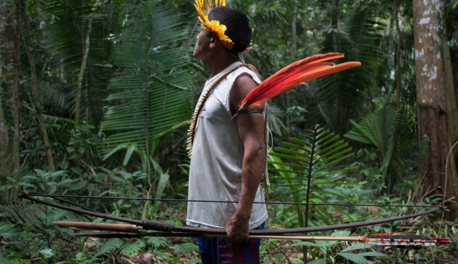 A man with wearing a crown of yellow feathers, a white tank top and shorts stands holding a bow and arrows in a densely wooded forest.