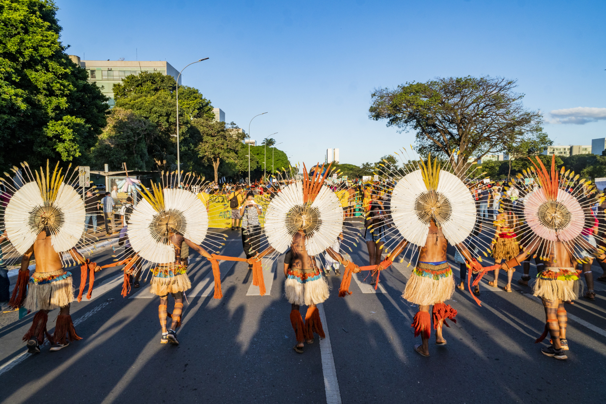 Five Indigenous activists wearing traditional dress march side by side in a demonstration on a tree-lined street on a sunny day.