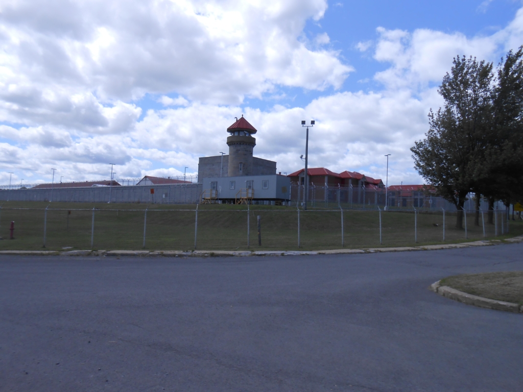 A complex of brick buildings with red rooves behind two rings of barbed-wire fence.