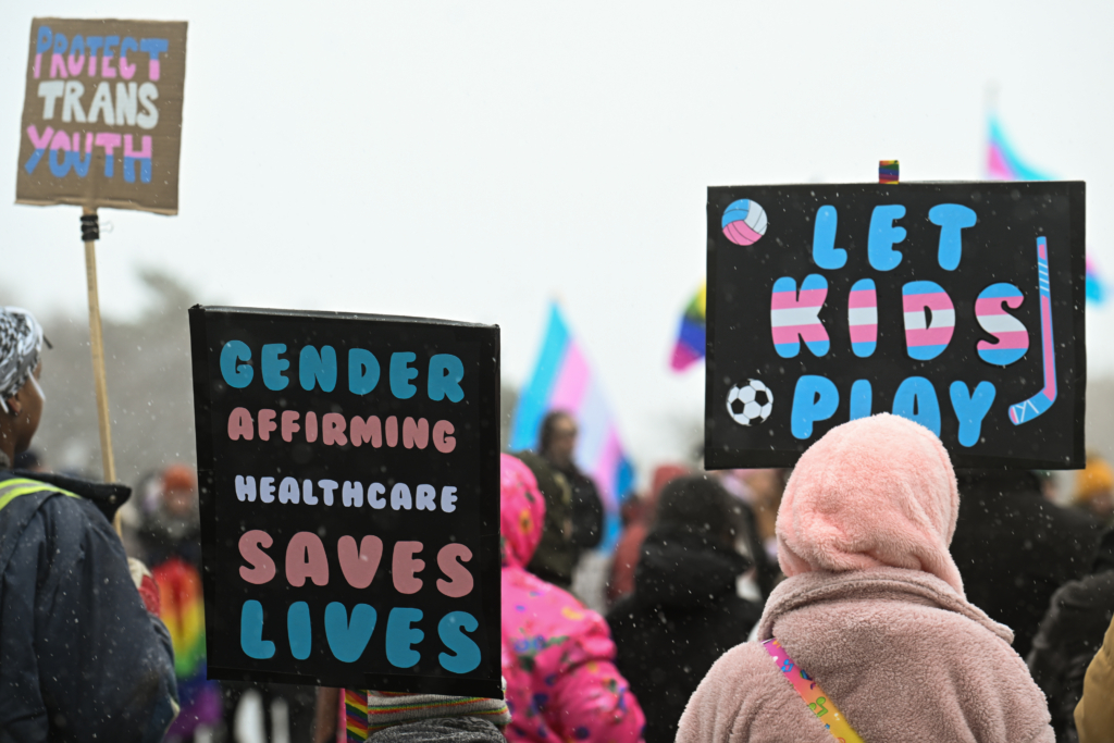 People at an outdoor demonstration hold protest signs that say "Gender-affirming care saves lives" and "Let kids play".