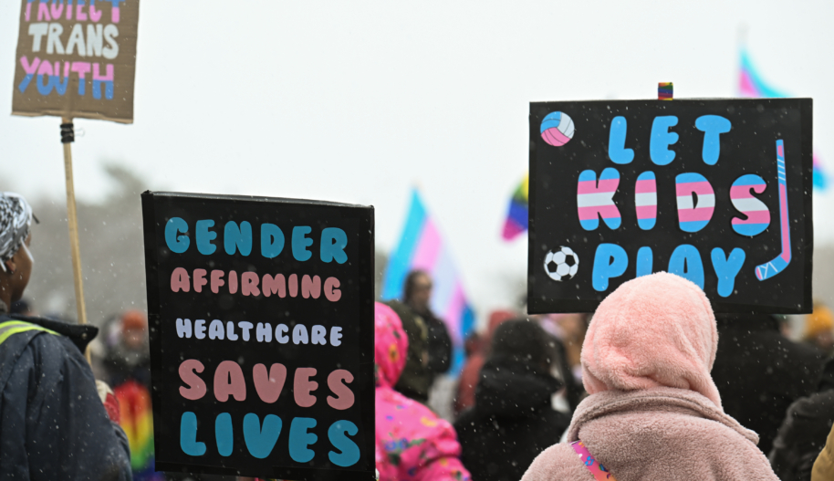 People at an outdoor demonstration hold protest signs that say "Gender-affirming care saves lives" and "Let kids play".