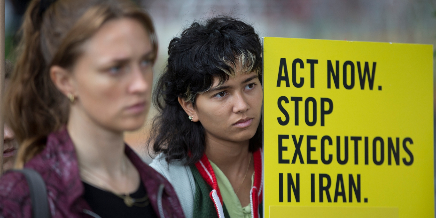 A woman with dark shoulder-length hair holds a yellow sign that reads "Act now, stop executions in Iran."