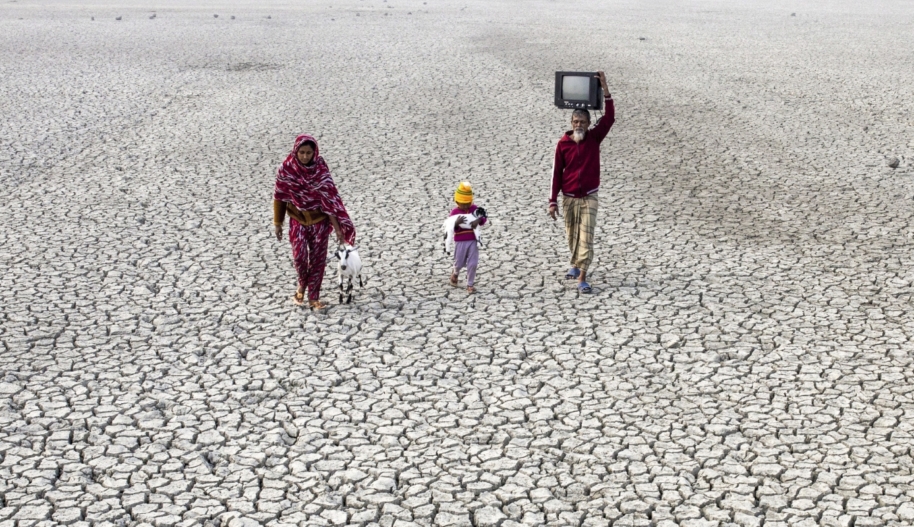 A child carrying a baby goats walks between a man carrying a TV on his head and a woman walking with an adult goat on soil parched by drought.