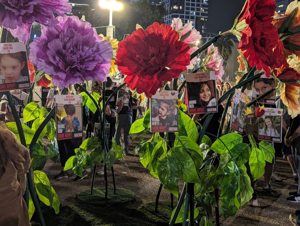 Header image: An installation in “Hostages Square” (official name “Tel Aviv Museum Plaza”), of adult human size flowers with signs of children hostages. Photo by Gil Naveh/Amnesty International Israel.