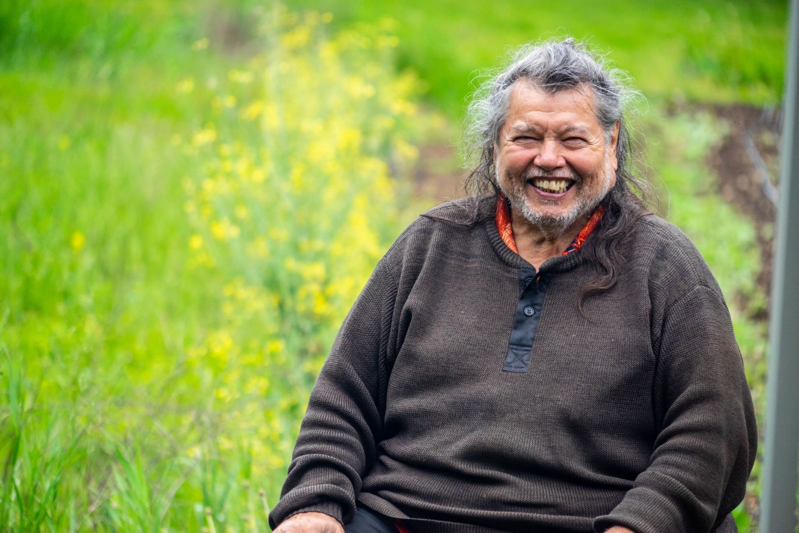 A man in a dark brown and long silver hair smiles while seated.