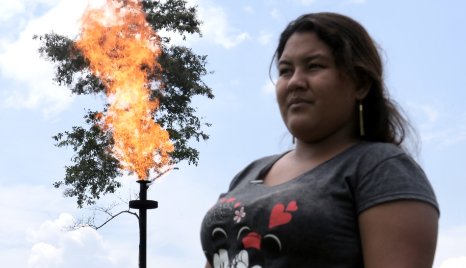 A young woman wearing a grey v-neck tshirt while a flame flares out of a pipe in the sky behind her.