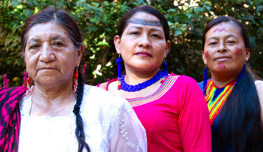 Three women Indigenous and environmental rights defenders pose outdoors in front of a garden