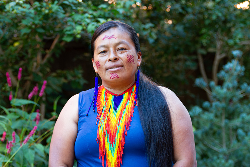 A woman with long black hair wearing a sleeveless blue top, long earrings with bright blue bead, and a necklace with blue, green, yellow, orange and red bead work. 