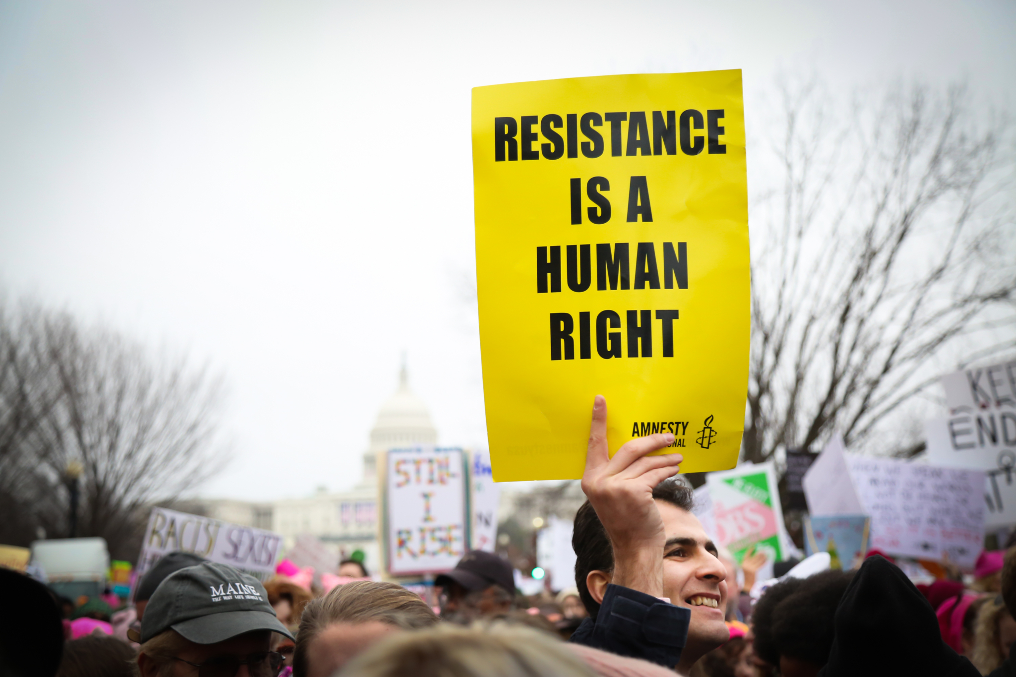 A man with grey and black hair at a demonstration holds a yellow sign with black text that reads "Resistance is a human right."