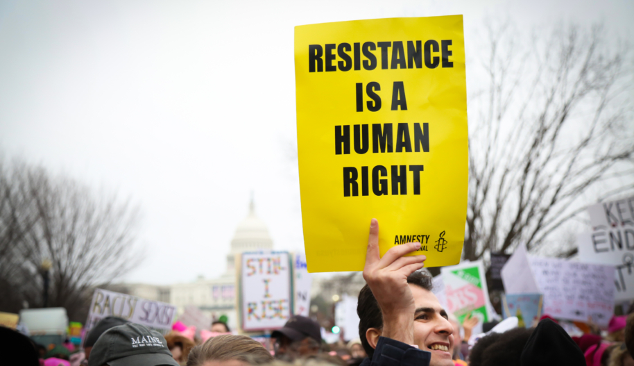 A man with grey and black hair at a demonstration holds a yellow sign with black text that reads "Resistance is a human right."