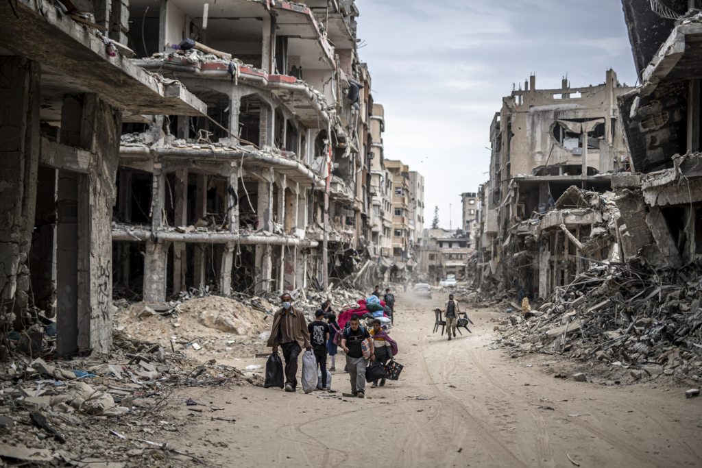 A group of men and youth carry furniture and other personal effects on a dirt road lined with bombed out buildings.