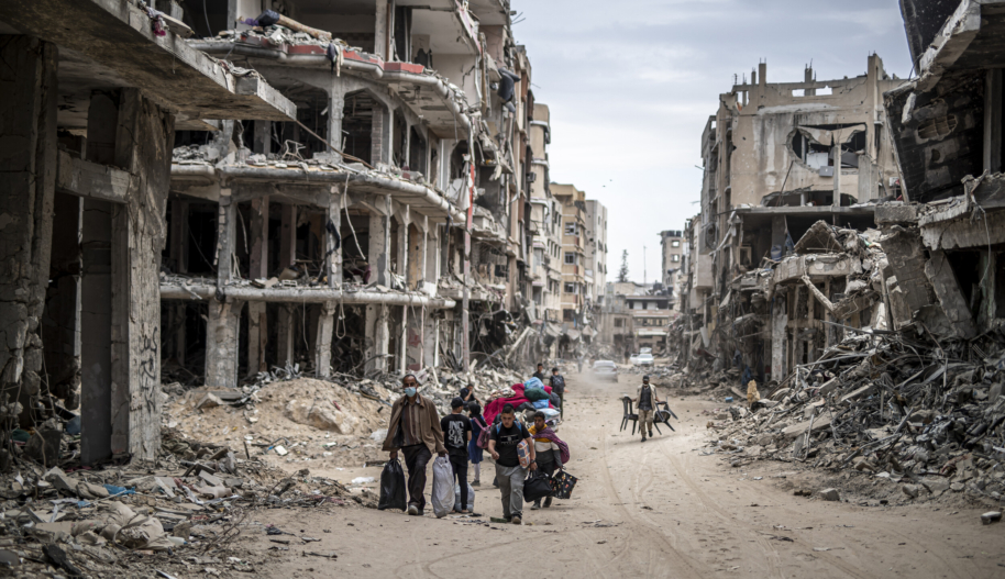 A group of men and youth carry furniture and other personal effects on a dirt road lined with bombed out buildings.