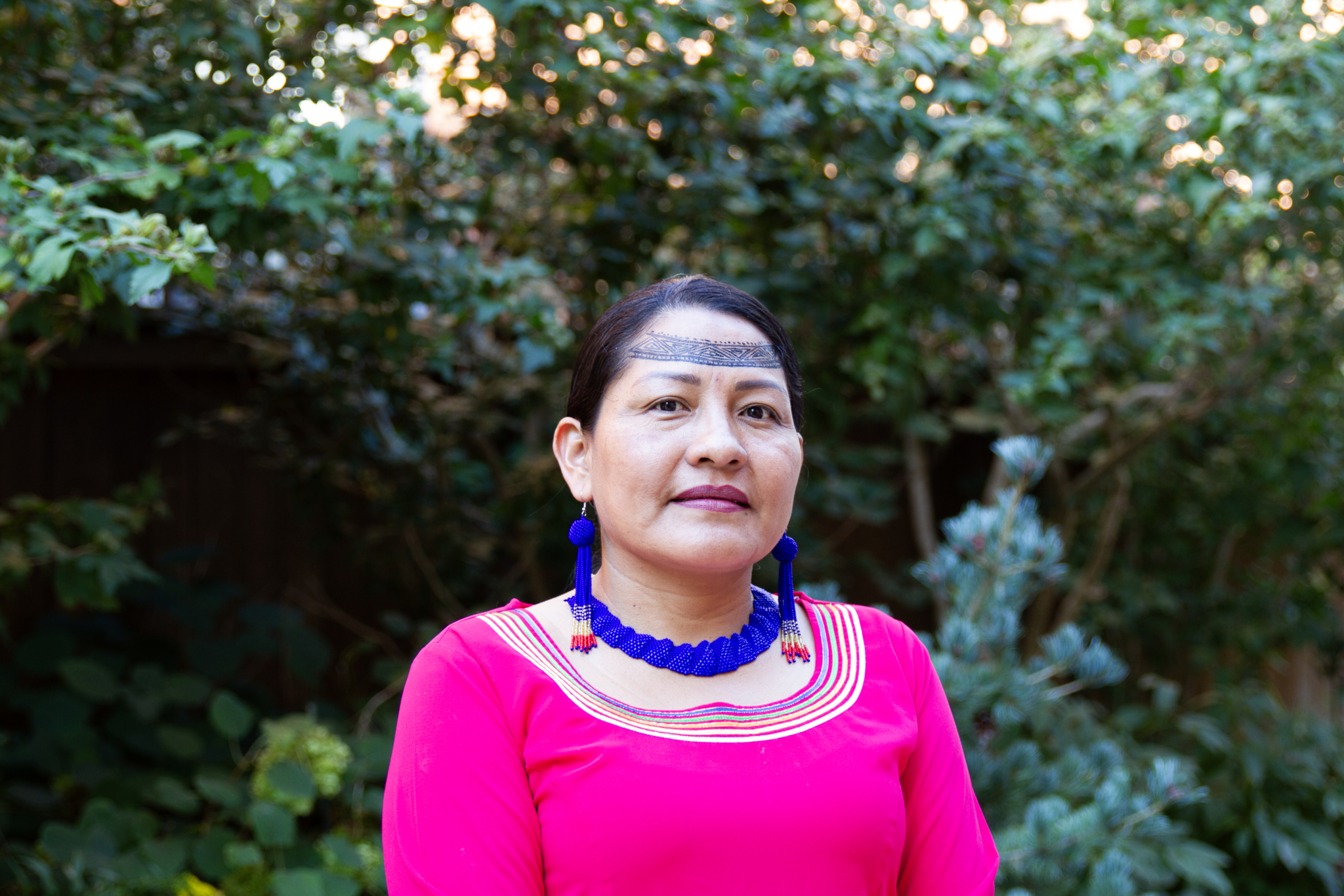 A woman wearing a bright pink long-sleeved shirt, a bright blue beaded necklace and bright blue beaded earrings.