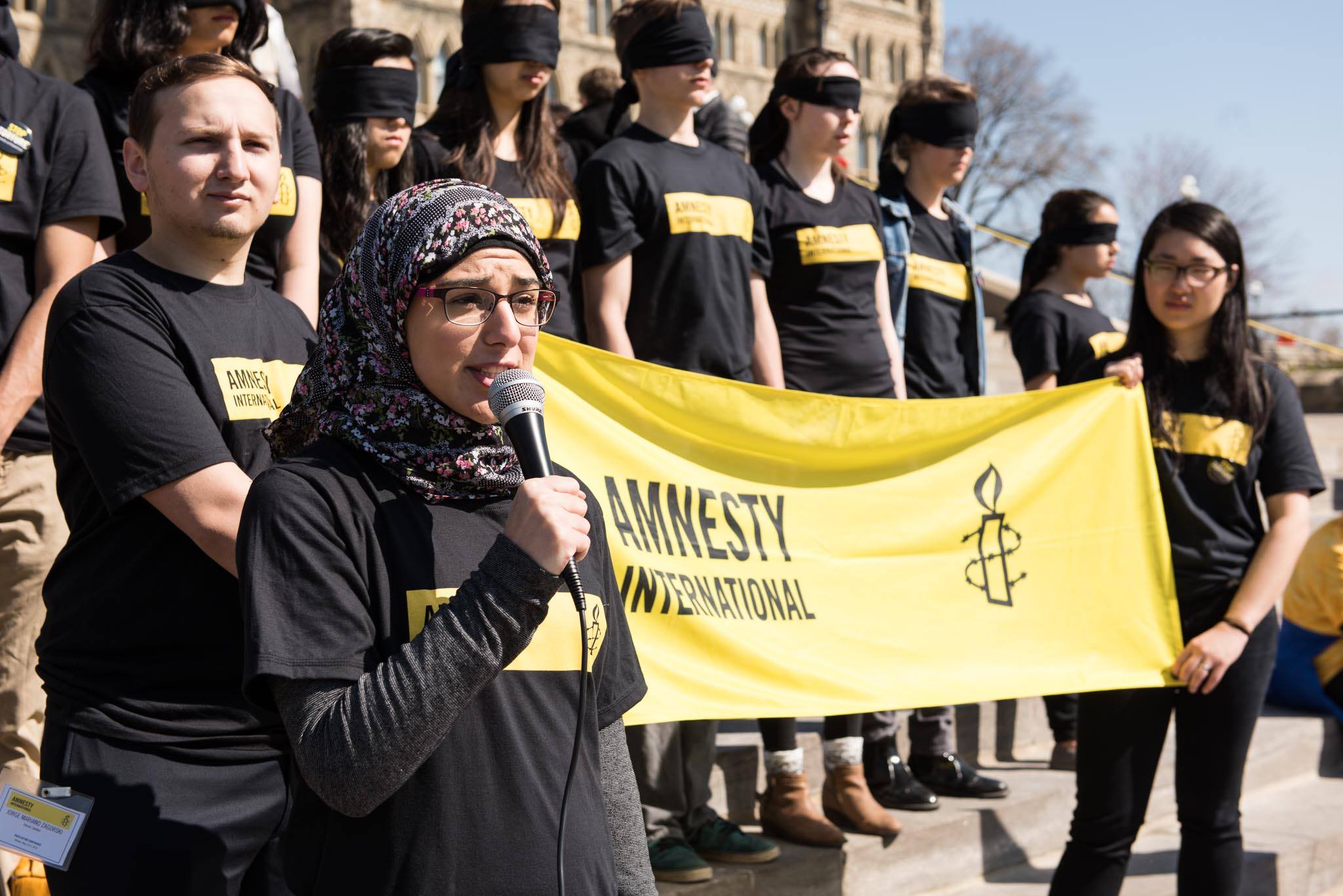 Amnesty supporters with their eyes covered and a women speaking into a microphone