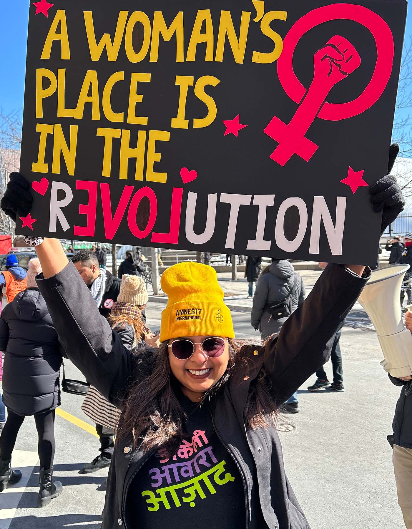 A woman smiling holding up a sign that reads "A Women's Place is in the Revolution"