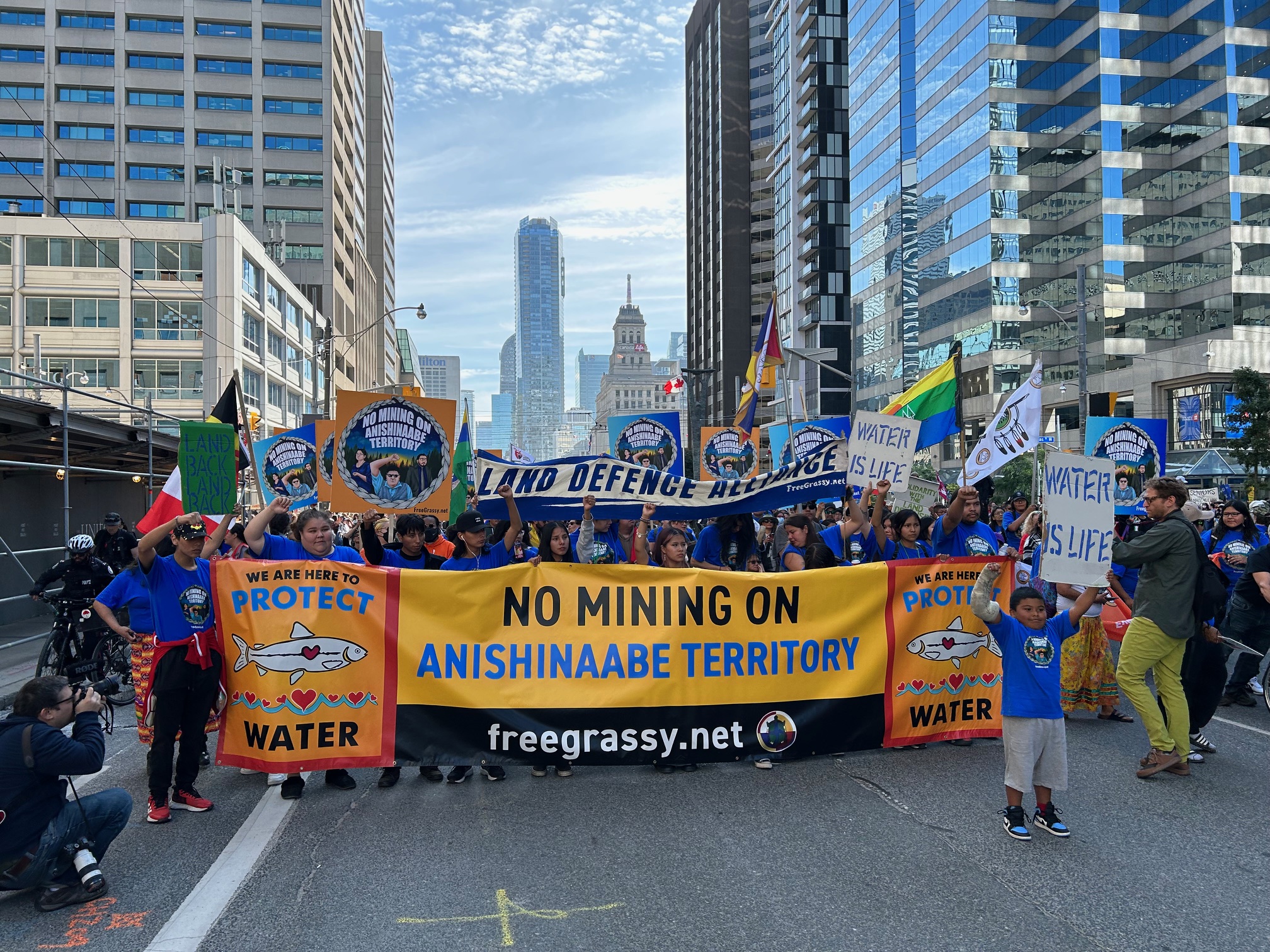 A group of protesters marches along University Avenue in downtown Toronto, holding signs and banners opposing mining in the Ring of Fire region. At the front, a yellow banner reads “No Mining on Anishinaabe Territory.” The demonstrators include people of diverse ages and backgrounds.