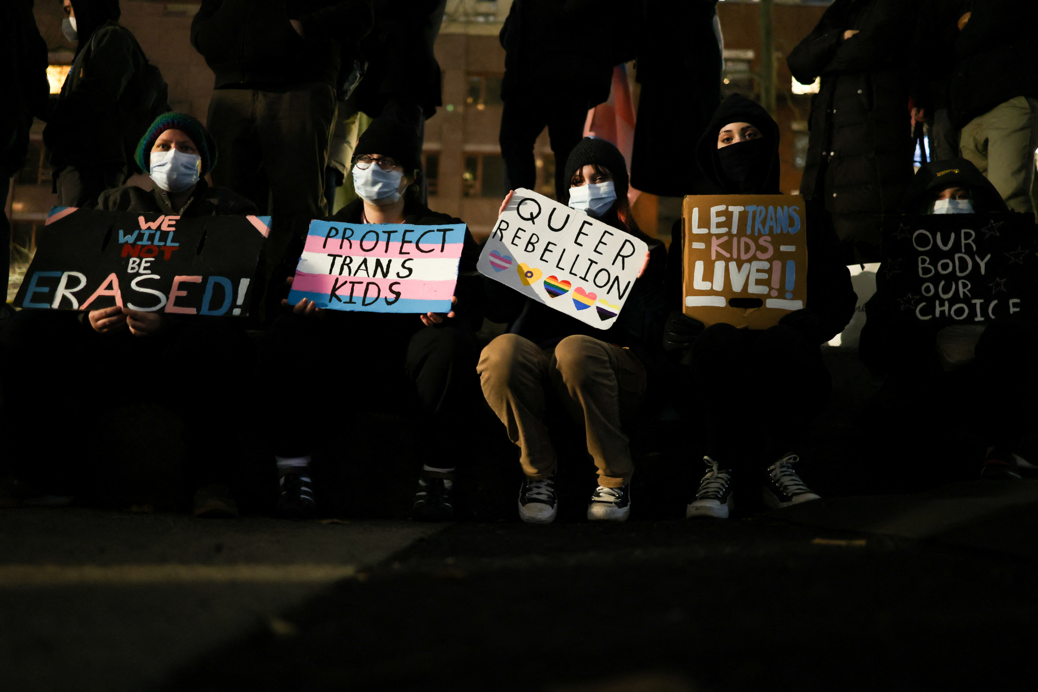 Three protesters sit in a row hold signs that read "Protect trans kid," "Queer rebellion," and "Let trans kids live," respectively.