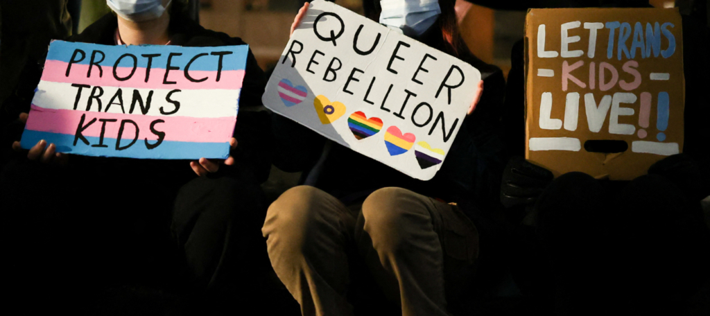 Three protesters sit in a row hold signs that read "Protect trans kid," "Queer rebellion," and "Let trans kids live," respectively.