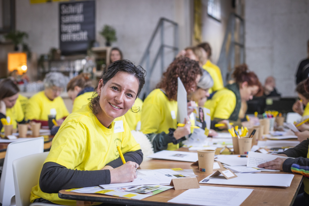 A young woman is seated at a long table with a pen in her hand and papers in front of her. She's wearing a yellow shirt with black long sleeves underneath. There are others at the table with her