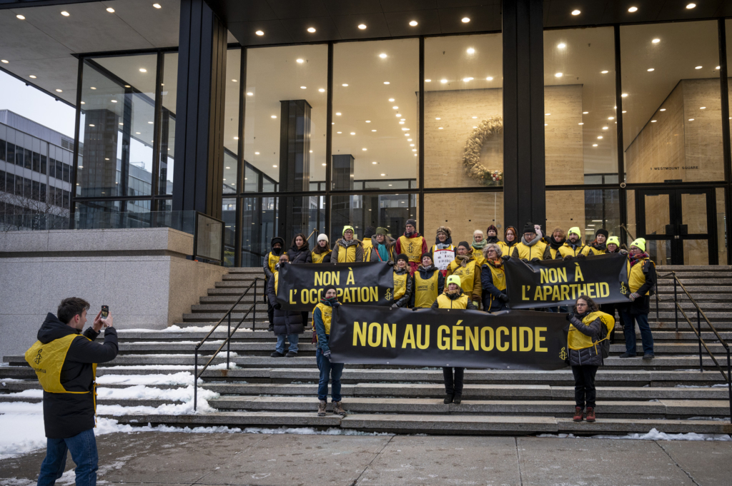 About 20 people stand on large concrete stairs in front of an office building holding black and yellow banners that read "No to genocide", "No to apartheid," and "No to occupation".
