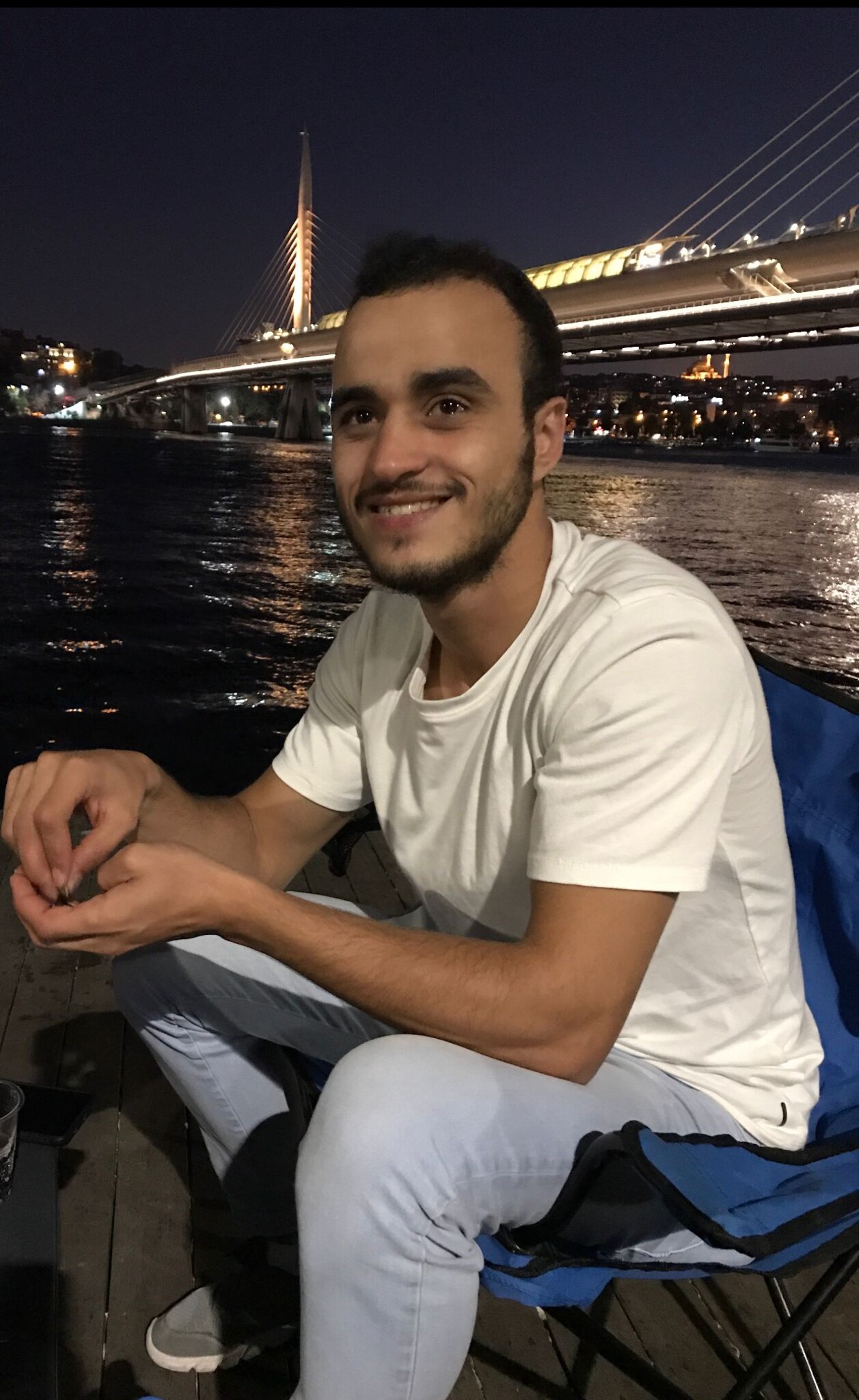 Ahmad Aabo wearing a white shirt and seated on a chair in front of a body of water. The picture appeared to have been taken at night and he is smiling into the camera