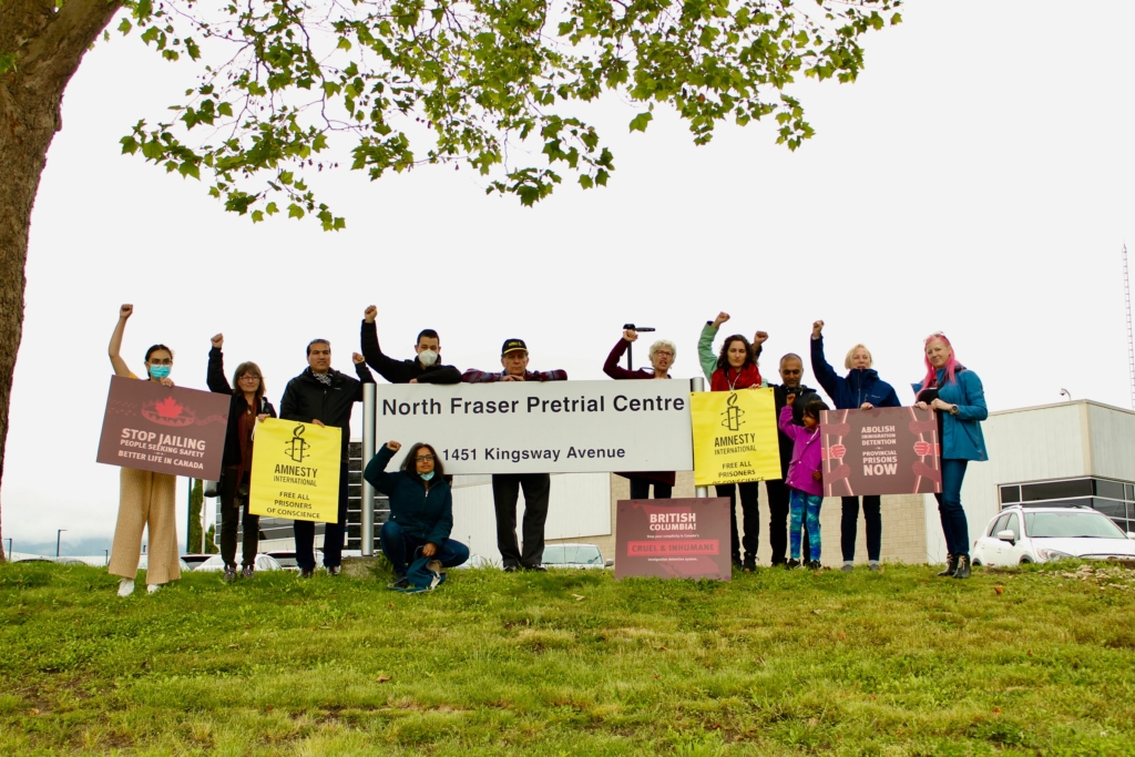 People holding signs calling for an end to immigration detention in Canada raise their fists at an outdoor rally in front of a jail.