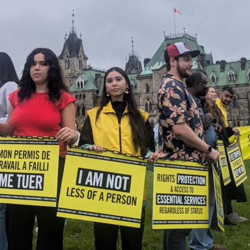 A group of about a dozen people stand on a lawn, holding yellow signs calling for justice for migrant workers in Canada.