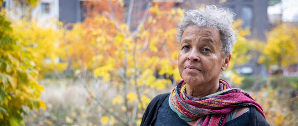 A woman with short, curly grey hair and a colourful scarf stands outside on an autumn day.