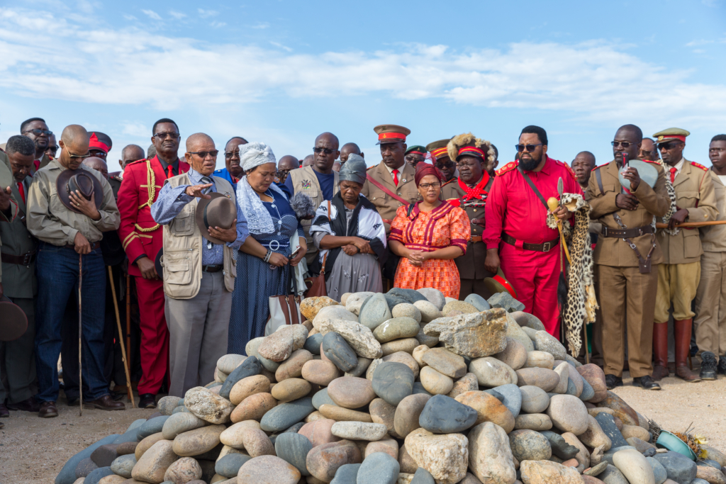 A group of people stand solemnly around a mound of smooth stones during a memorial ceremony, some with heads bowed and hats removed, under a bright blue sky.