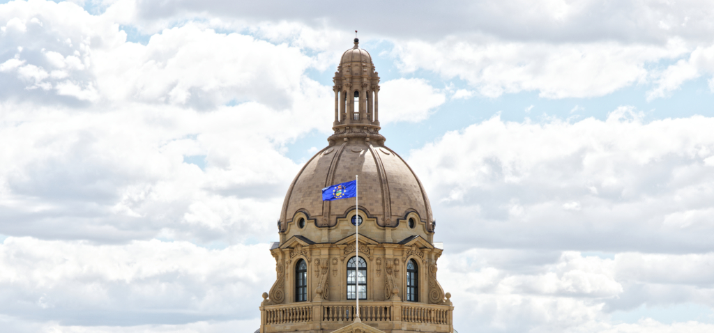 A sunny view of the Alberta Legislature Building in Edmonton, showing its domed sandstone façade reflected in a pool, with trees and a blue sky in the background.