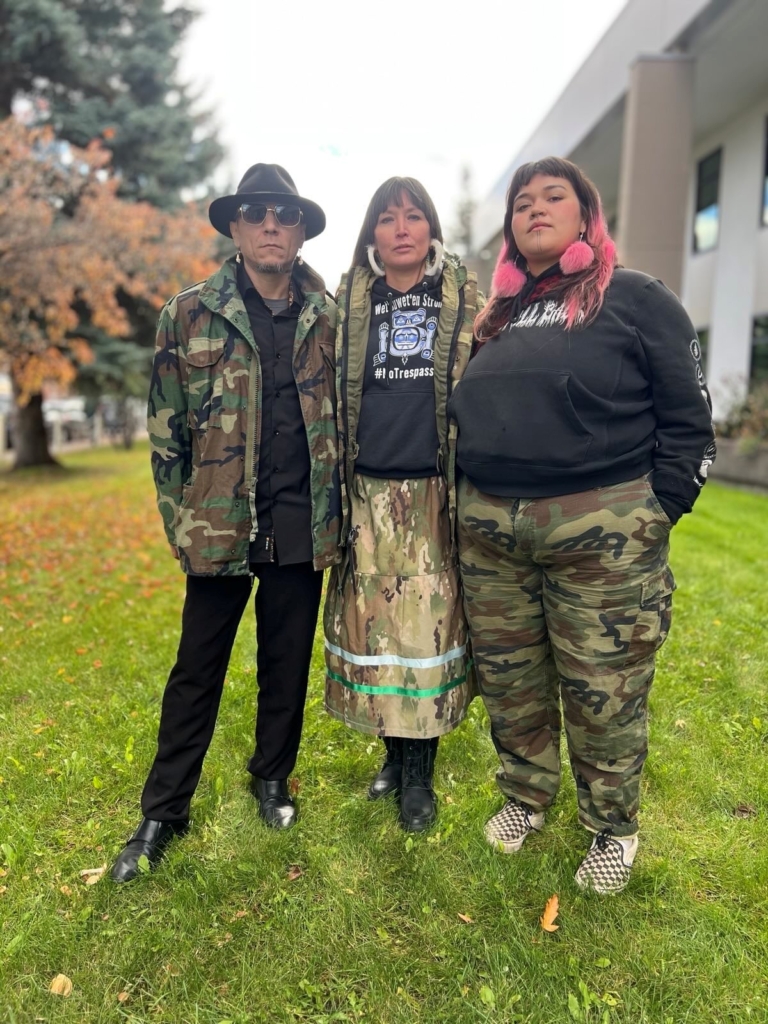 A man, a woman, and another woman stand in a row outside on an overcast day.