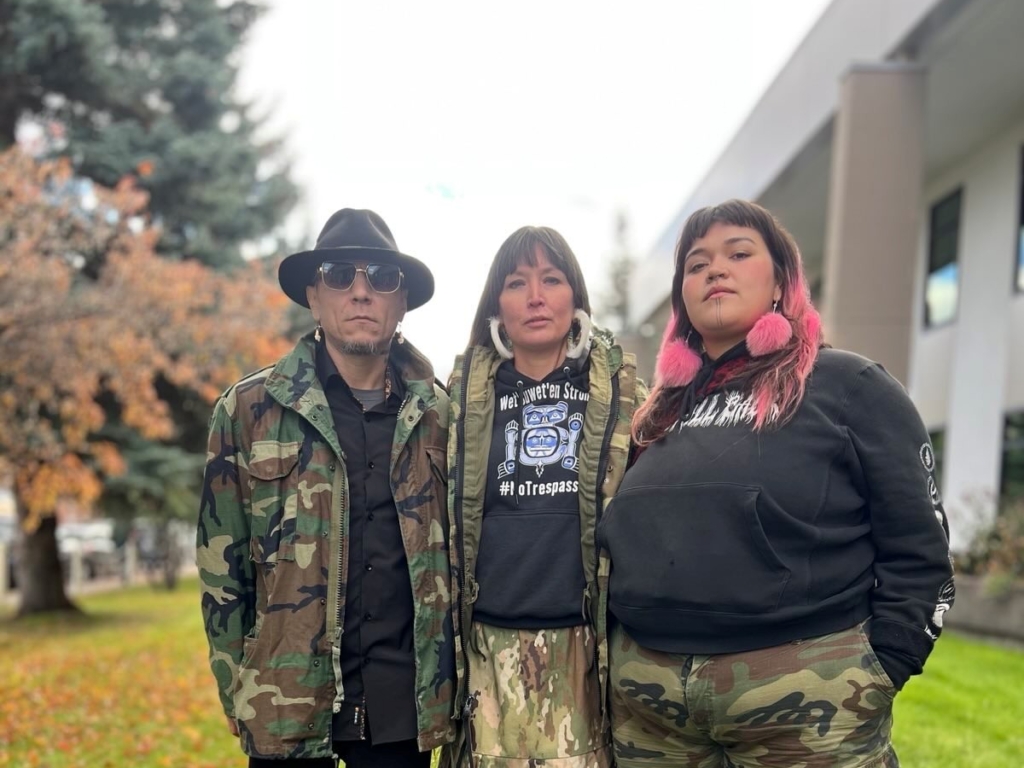 A man, a woman, and another woman stand in a row outside on an overcast day.