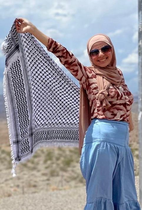Leqaa is pictured outside wearing a a brown long sleeve blouse and a hijab. She's smiling into the camera whilst holding a scarf
