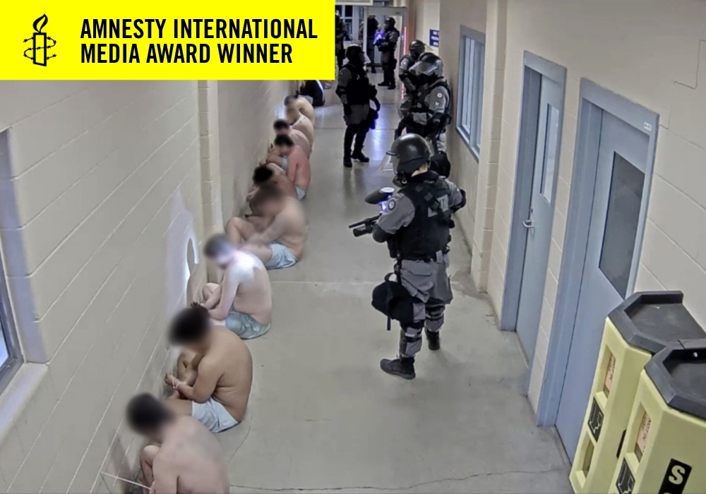 Officers in jail hallway aiming guns at prisoners who are on the ground facing the wall with only underwear on.