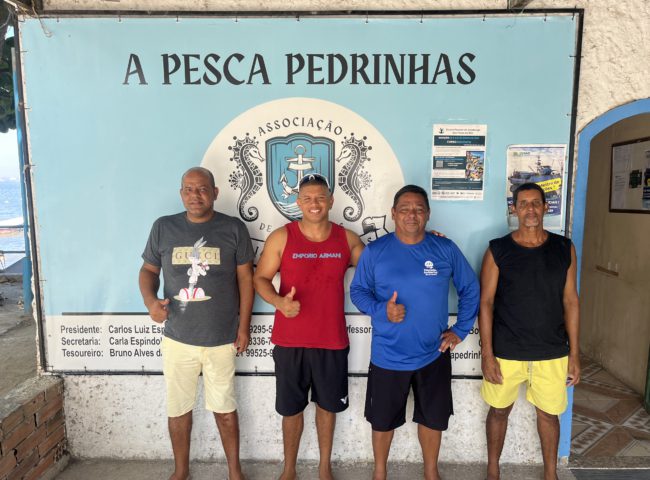 A row of four men in shorts stand with thumbs up in front of a sign which reads 'a pesca pedrinhas'.