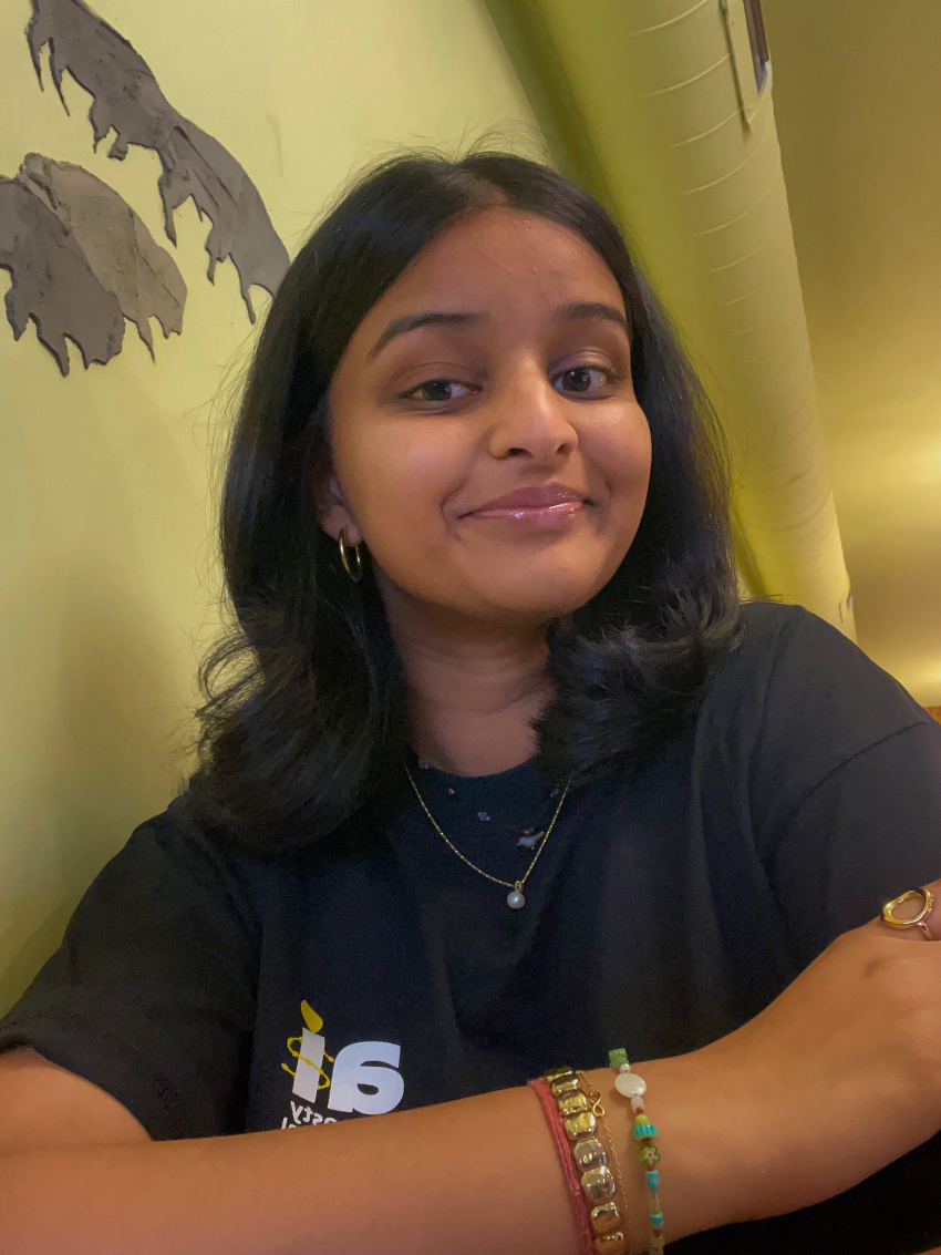 Veda Chandra wearing a black t-shirt with her arm folded on her chest and smiling into the camera.