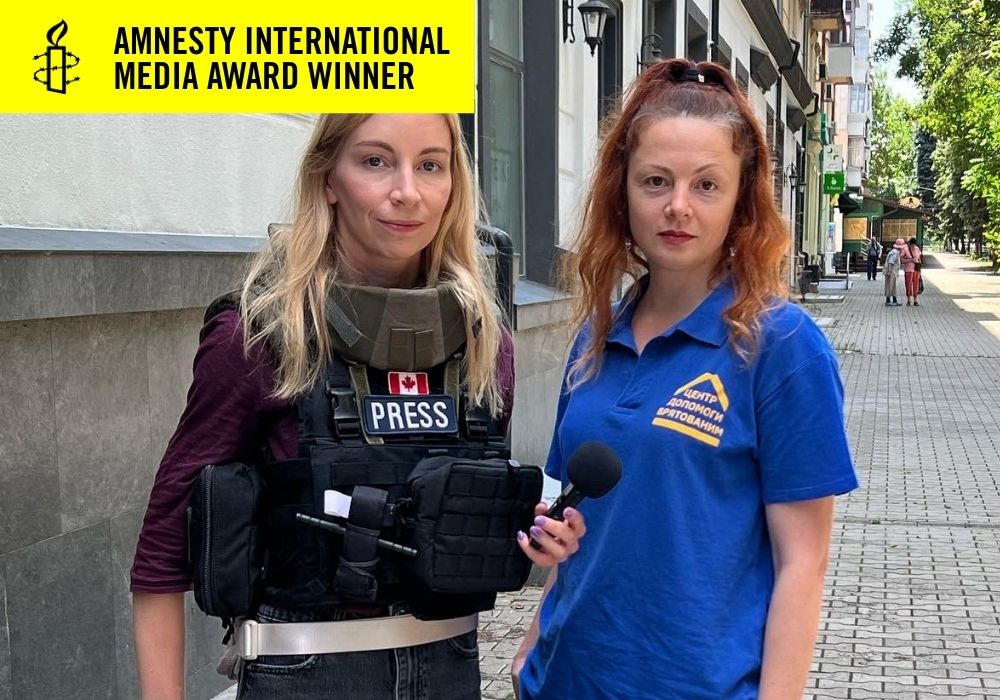 2 women standing on a sidewalk, one in gear with a "Press" label and the other in a blue company t-shirt